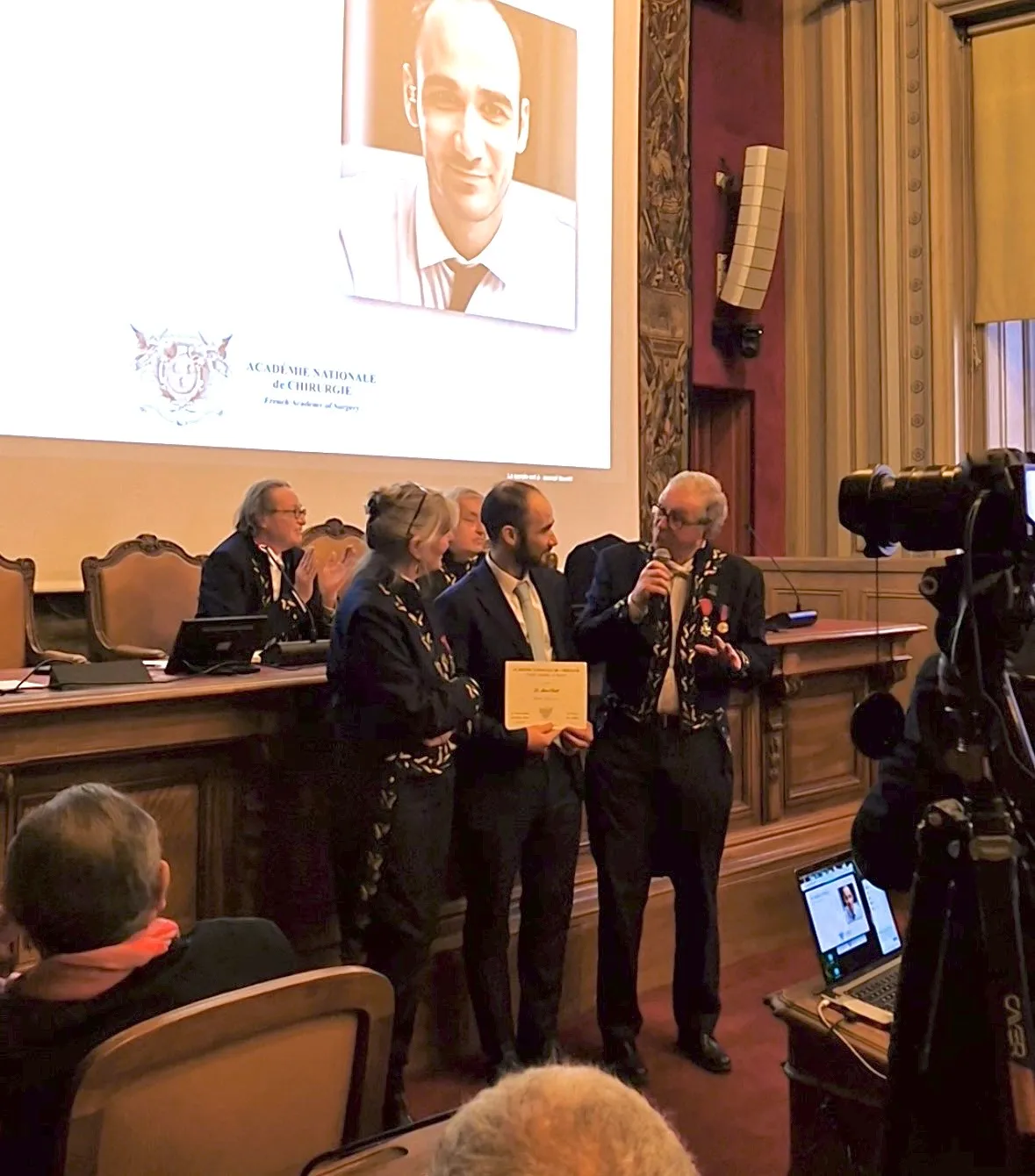 Le Dr Chelli reçoit son diplôme de membre de l'Académie nationale de chirurgie, entouré de deux académiciens en habit, devant un écran affichant son portrait et le logo de l'Académie.